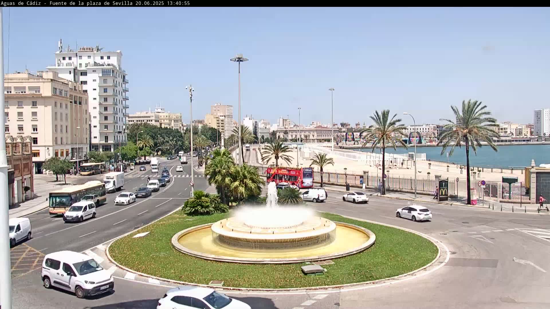 A city roundabout with a fountain in the center, surrounded by traffic and buildings, overlooks a waterfront area on a sunny day.