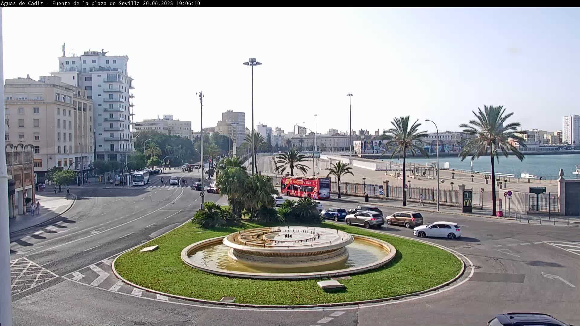 A circular fountain sits in a traffic circle near a waterfront, with buildings, palm trees, and a red double-decker bus visible under a bright, sunny sky.