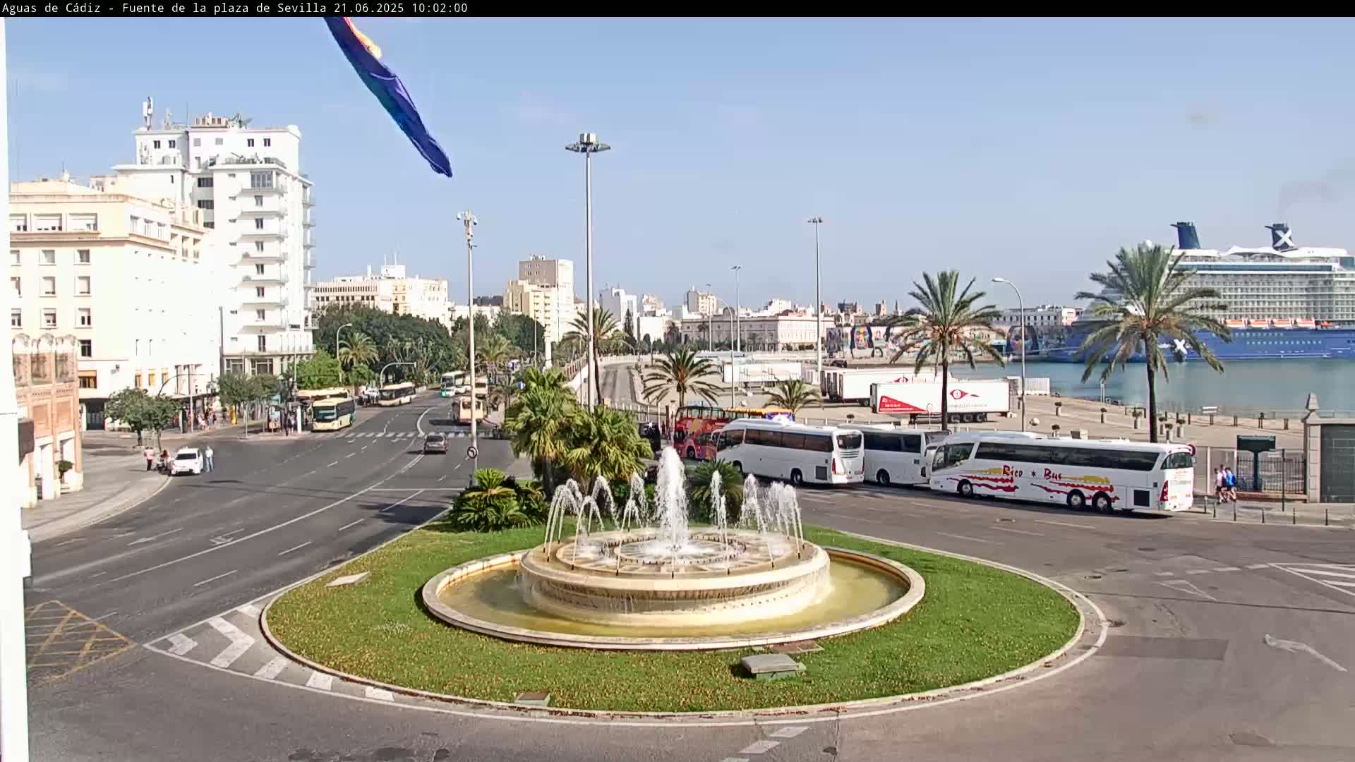 A circular fountain in a city square is surrounded by roads with buses and cars, and in the background, a cruise ship is docked near buildings under a clear sky.