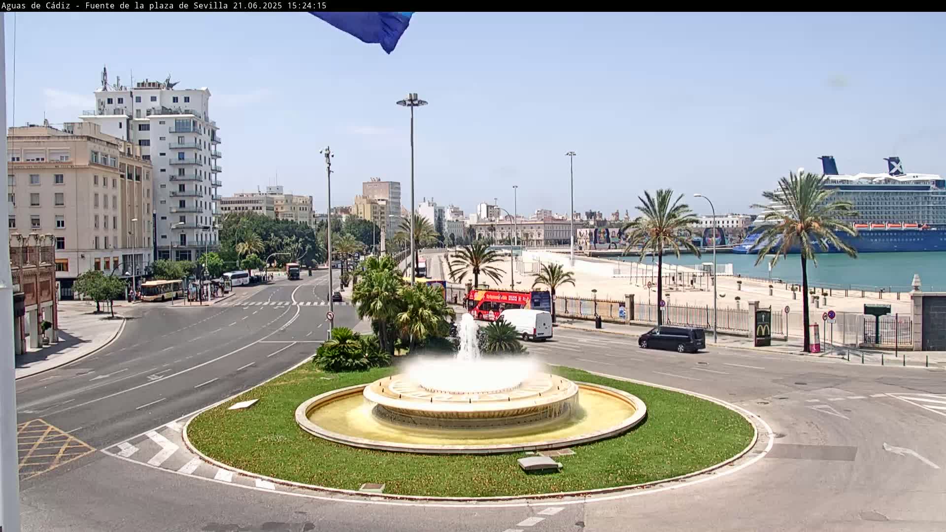 A circular fountain is at the center of a city roundabout near a harbor where a large cruise ship is docked, under a clear sunny sky.