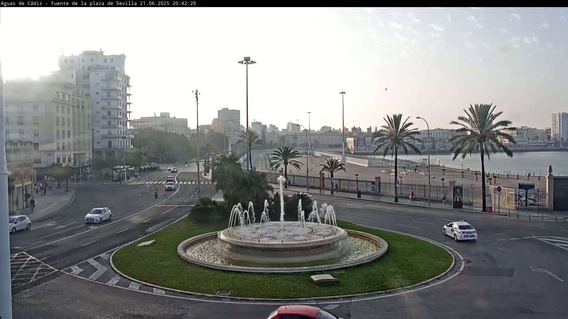 A roundabout with a central fountain is surrounded by city streets, buildings, and palm trees on a partly cloudy day.