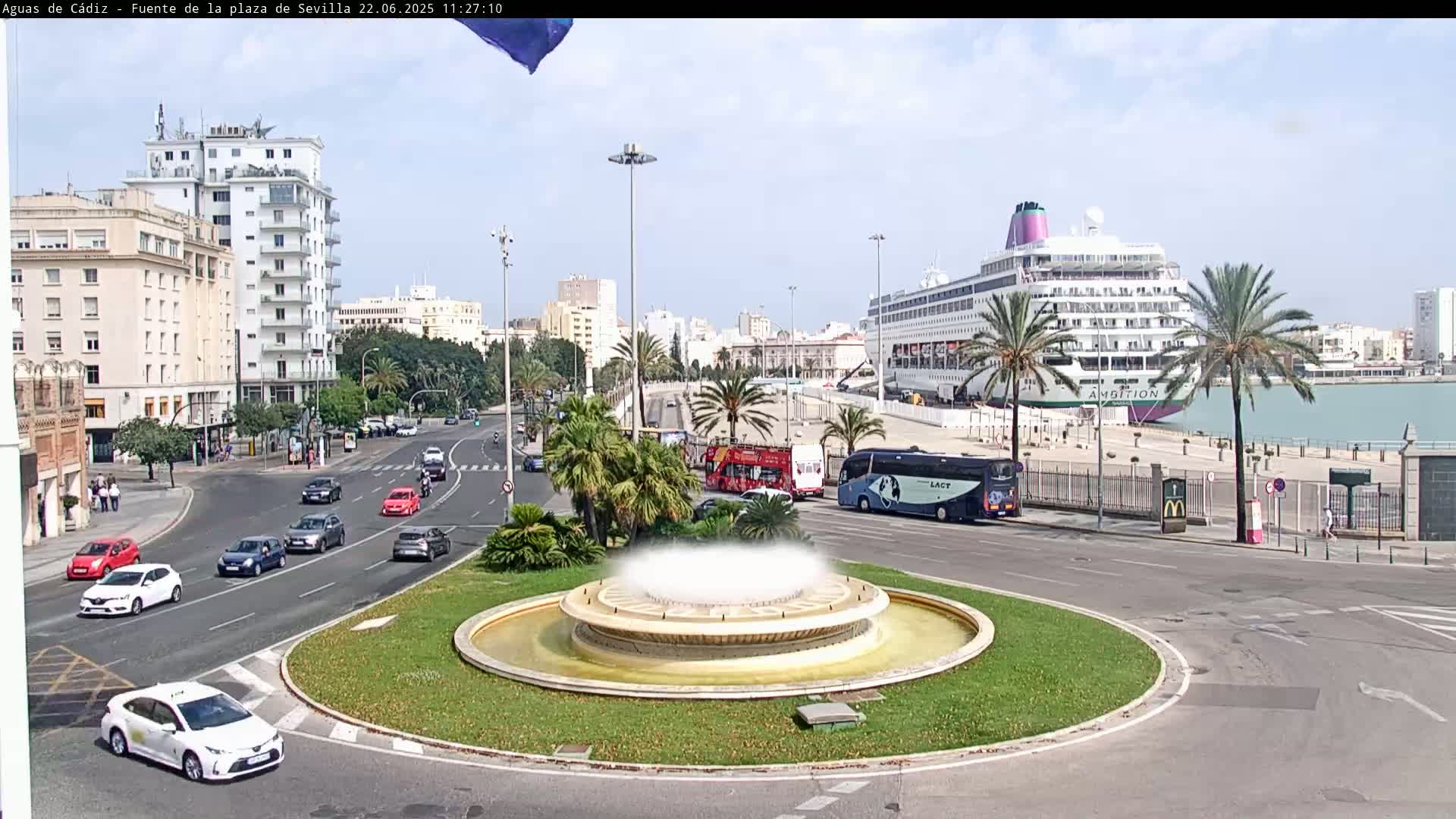 A city roundabout with a fountain in the center features cars driving on the road, a large cruise ship docked in the background, and palm trees lining the roadway under a clear sky.