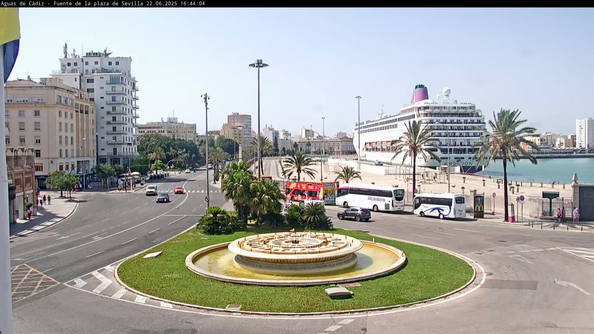 A city roundabout with a central fountain is shown, with several cars and tour buses nearby, and a large cruise ship docked in the harbor in the background under a clear sky.