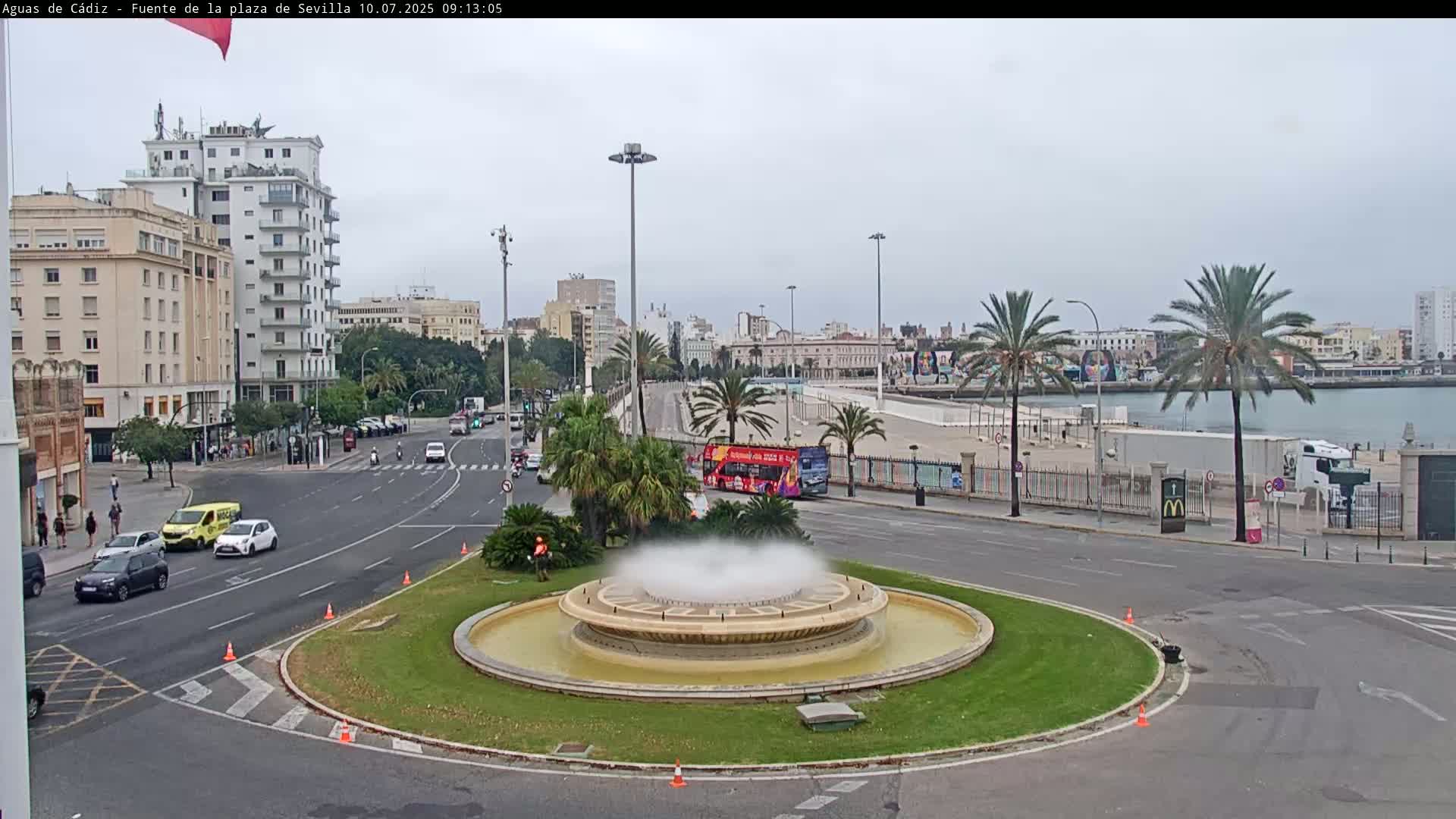 A city square with a fountain in the center, surrounded by roads with cars and a large building in the background under an overcast sky.
