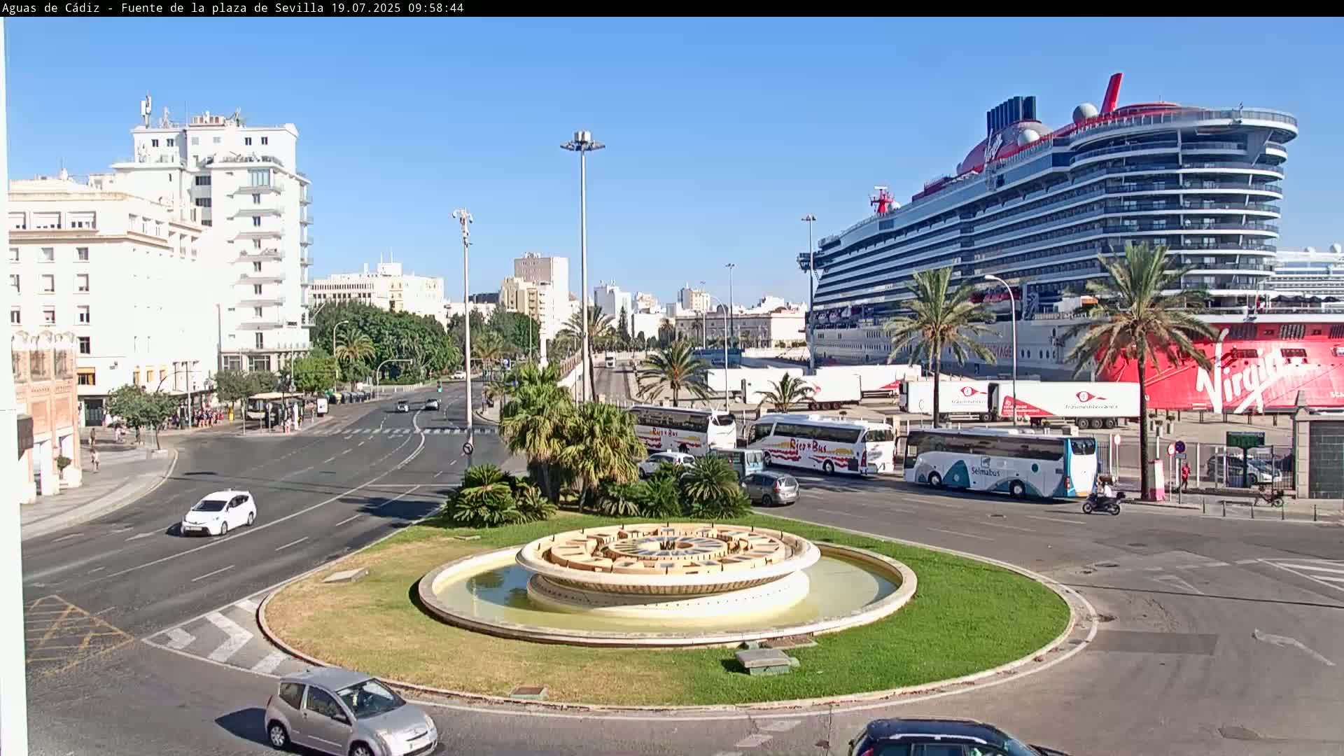 A large cruise ship is docked near a roundabout with a fountain, several buses, and cars; the weather is sunny.