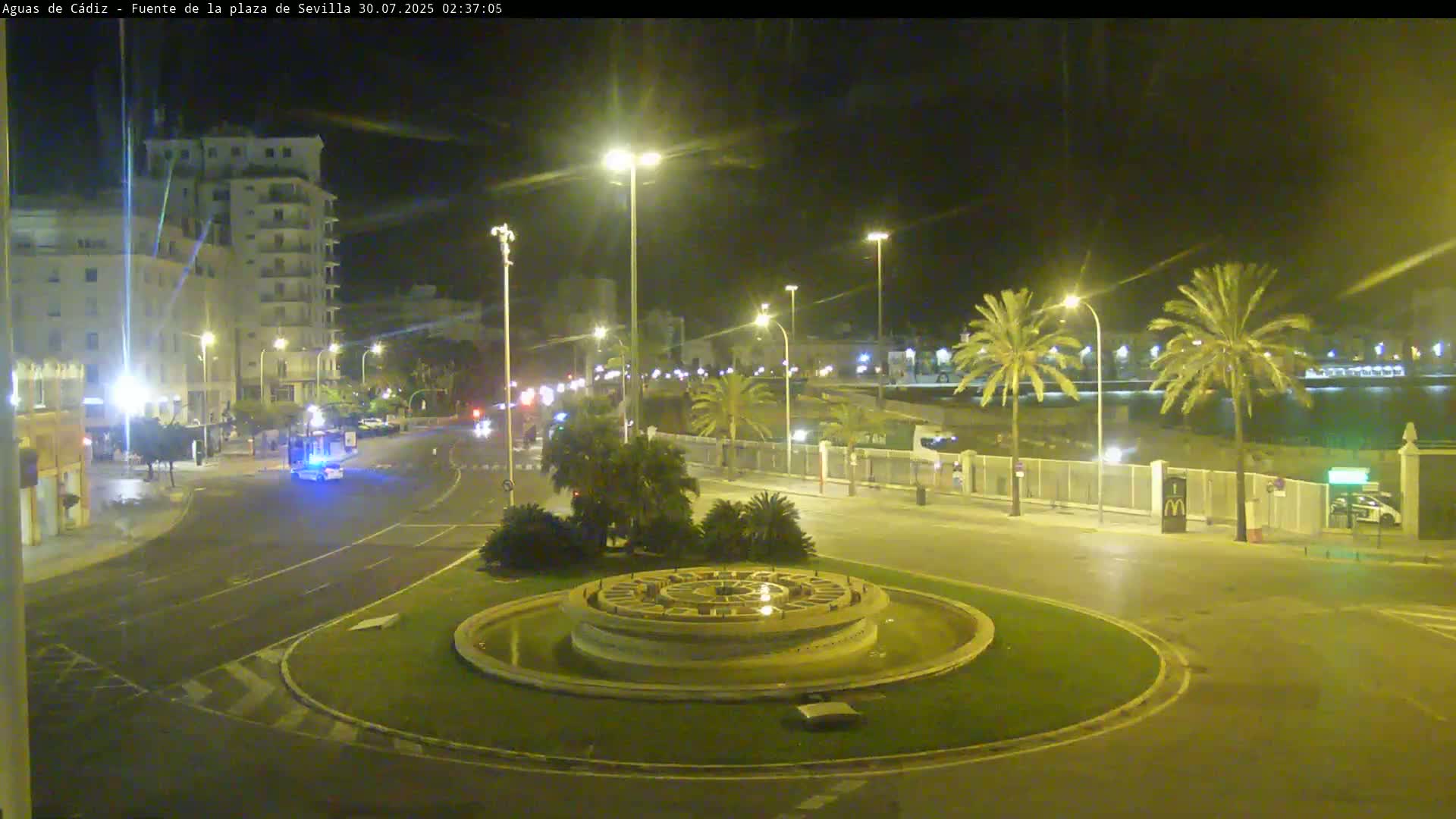 A large cruise ship is docked near a roundabout with a fountain, several buses, and cars; the weather is sunny.