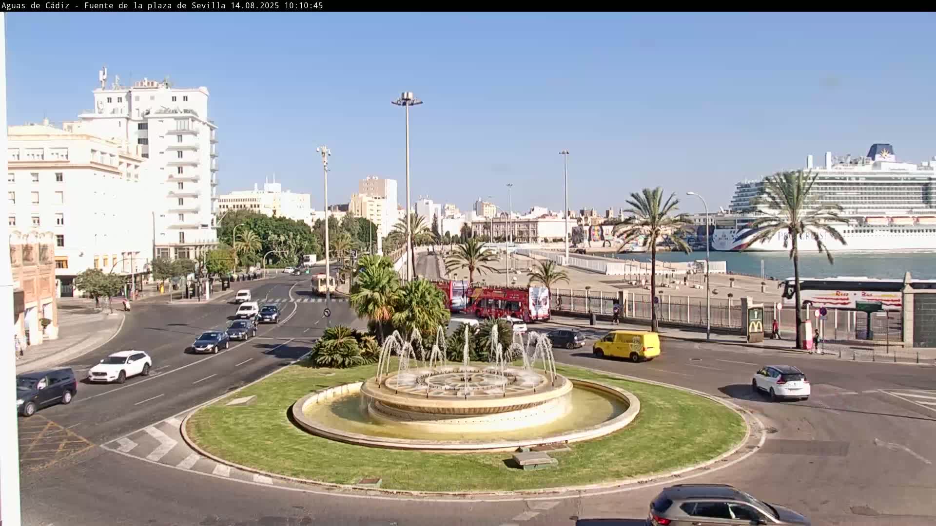 A circular fountain in a traffic circle is surrounded by cars, palm trees, and buildings, with a large cruise ship visible in the harbor under a clear, sunny sky.