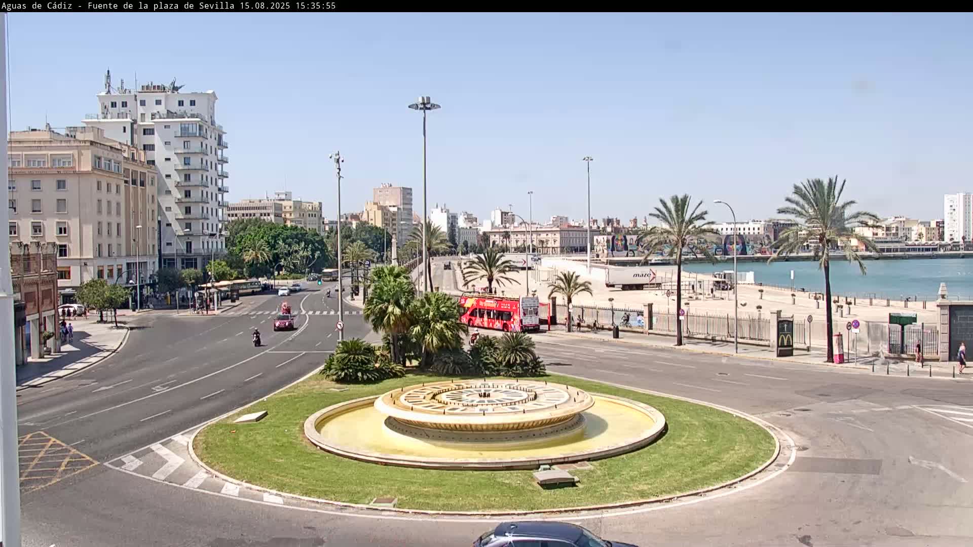 A circular fountain in a traffic circle is surrounded by cars, palm trees, and buildings, with a large cruise ship visible in the harbor under a clear, sunny sky.