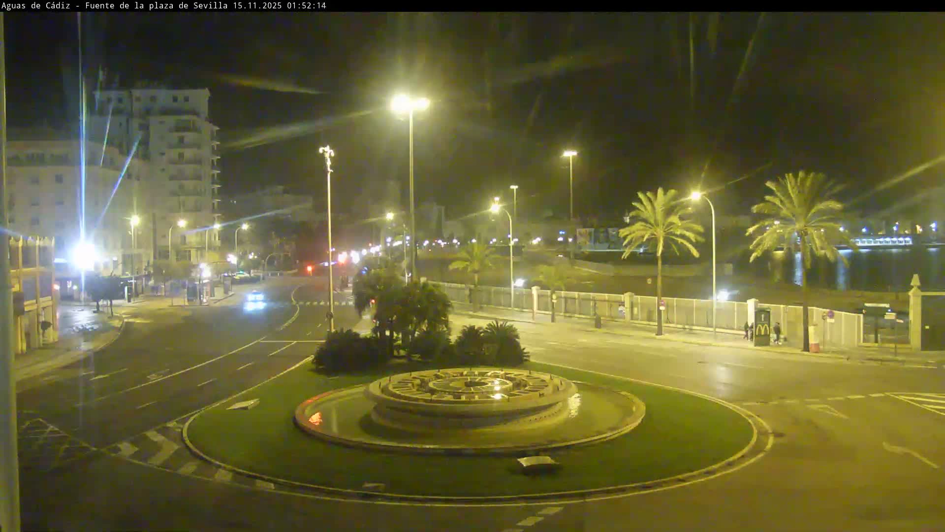 An urban intersection at night features a central decorative fountain roundabout, surrounded by illuminated buildings, palm trees, and sparse vehicle traffic under a clear sky.