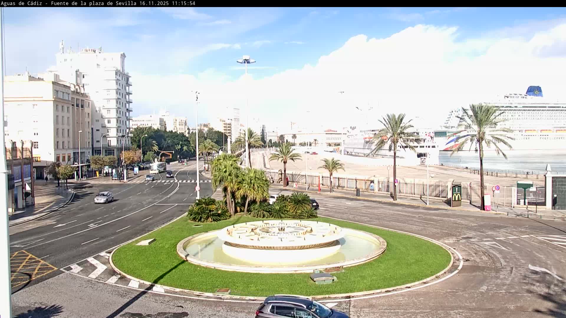 Under a bright, sunny sky with scattered clouds, a large city traffic circle featuring a decorative fountain is flanked by multi-story buildings and busy roads, leading towards a port where two cruise ships are docked among palm trees.