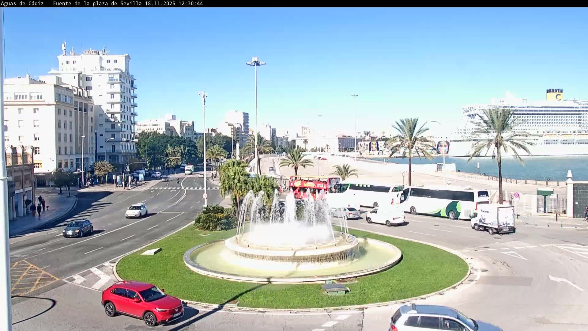 Under a bright, sunny sky with scattered clouds, a large city traffic circle featuring a decorative fountain is flanked by multi-story buildings and busy roads, leading towards a port where two cruise ships are docked among palm trees.