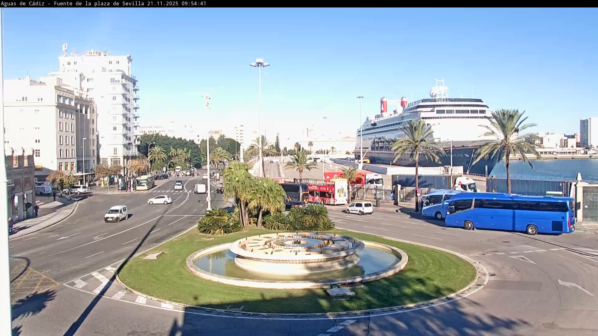 On a clear, sunny day, a bustling city street with cars and buses curves around a central fountain, leading towards a port where a large cruise ship is docked, flanked by palm trees and buildings.