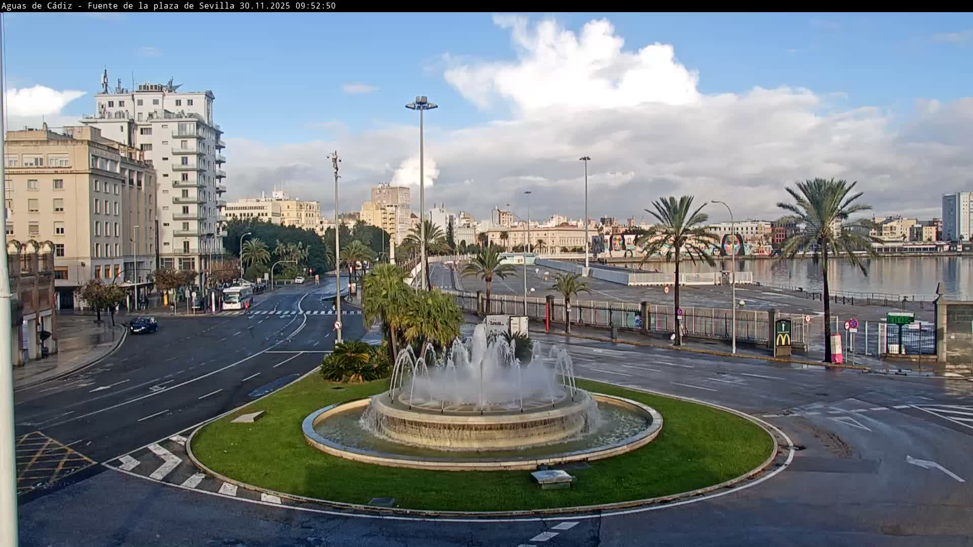 A city square with a prominent fountain in a grassy roundabout is surrounded by wet roads, buildings, and palm trees, with a waterfront visible in the background under a partly cloudy sky.