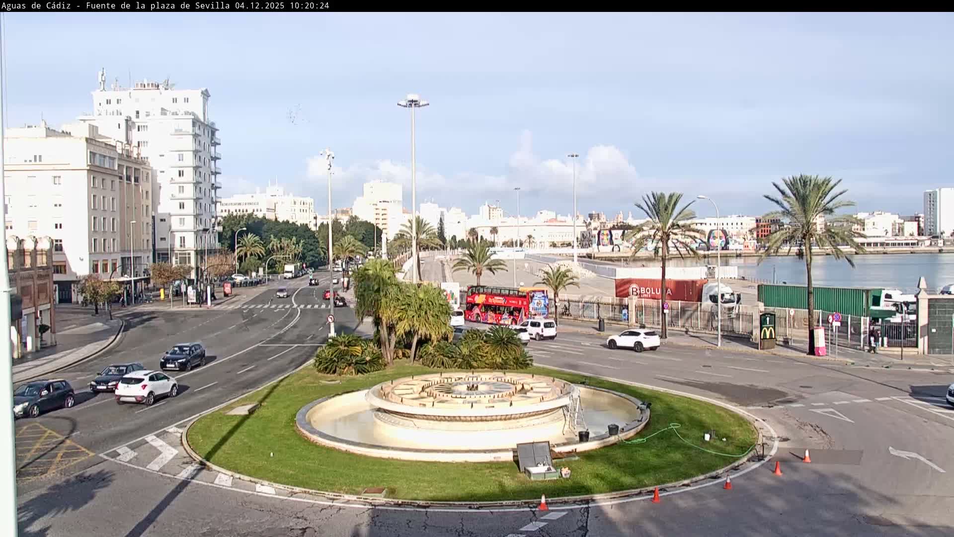 On a sunny, partly cloudy day, a city view captures a large traffic roundabout with a central fountain, surrounded by urban buildings, numerous palm trees, and active roadways, all with a harbor and industrial containers visible in the background.