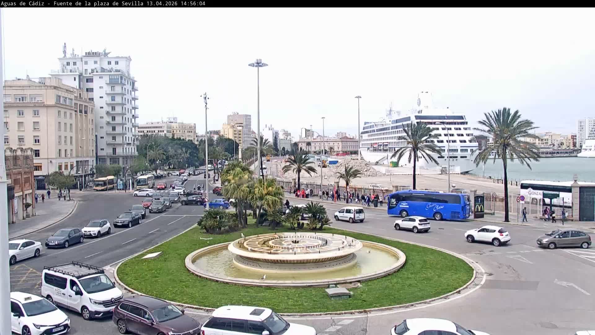 On a sunny, partly cloudy day, a city view captures a large traffic roundabout with a central fountain, surrounded by urban buildings, numerous palm trees, and active roadways, all with a harbor and industrial containers visible in the background.