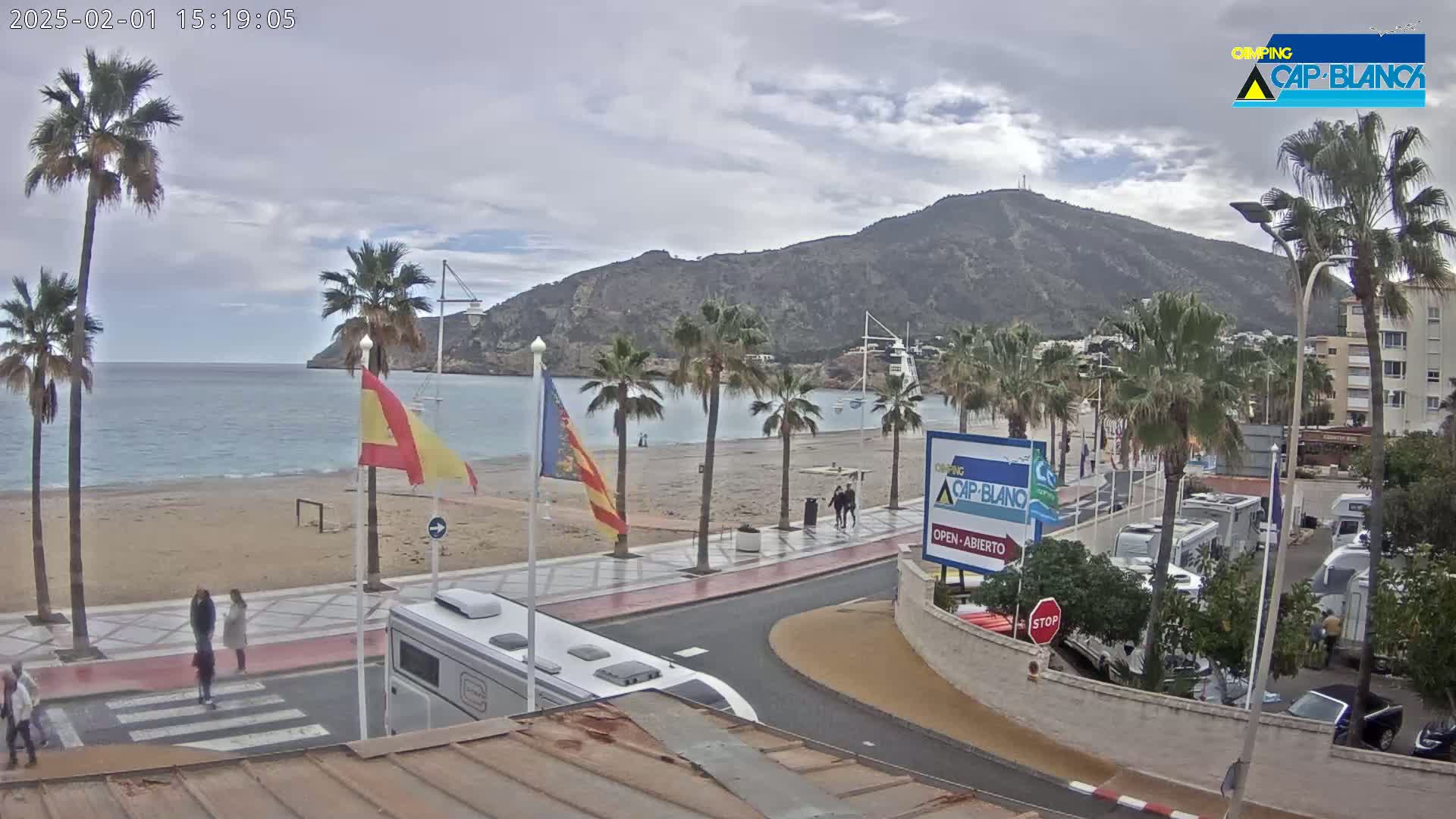 A beach with palm trees and a seaside promenade is visible under an overcast sky, with a mountain in the background and several RVs parked nearby.