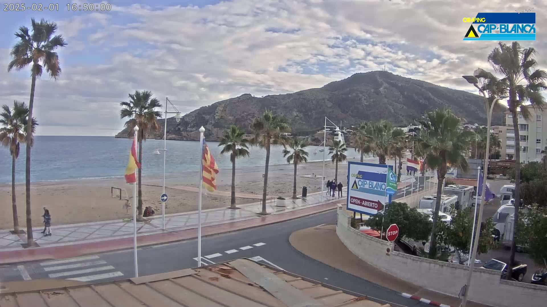 A sandy beach with palm trees lines a paved walkway next to calm ocean water under a partly cloudy sky, with a mountain in the background.
