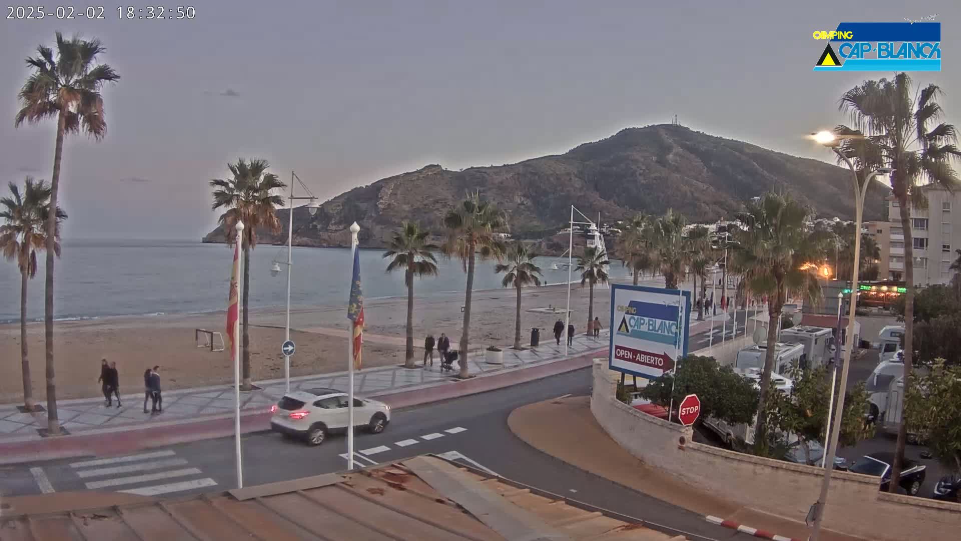 A seaside town at dusk shows a sandy beach, palm trees, a paved walkway, a few pedestrians, and vehicles parked near buildings at the foot of a hill.