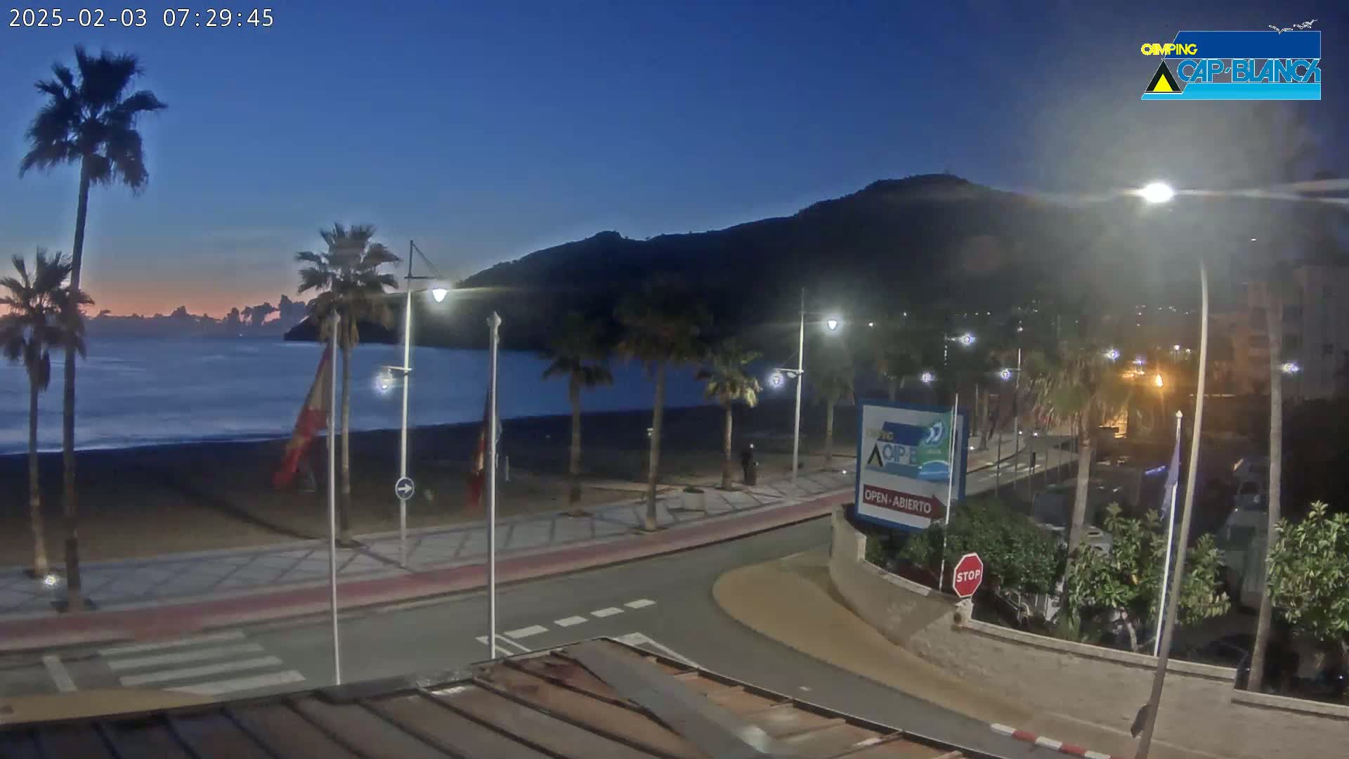 A mostly dark, beachside promenade with palm trees and streetlights under a twilight sky, overlooking a calm ocean and a hill in the background.