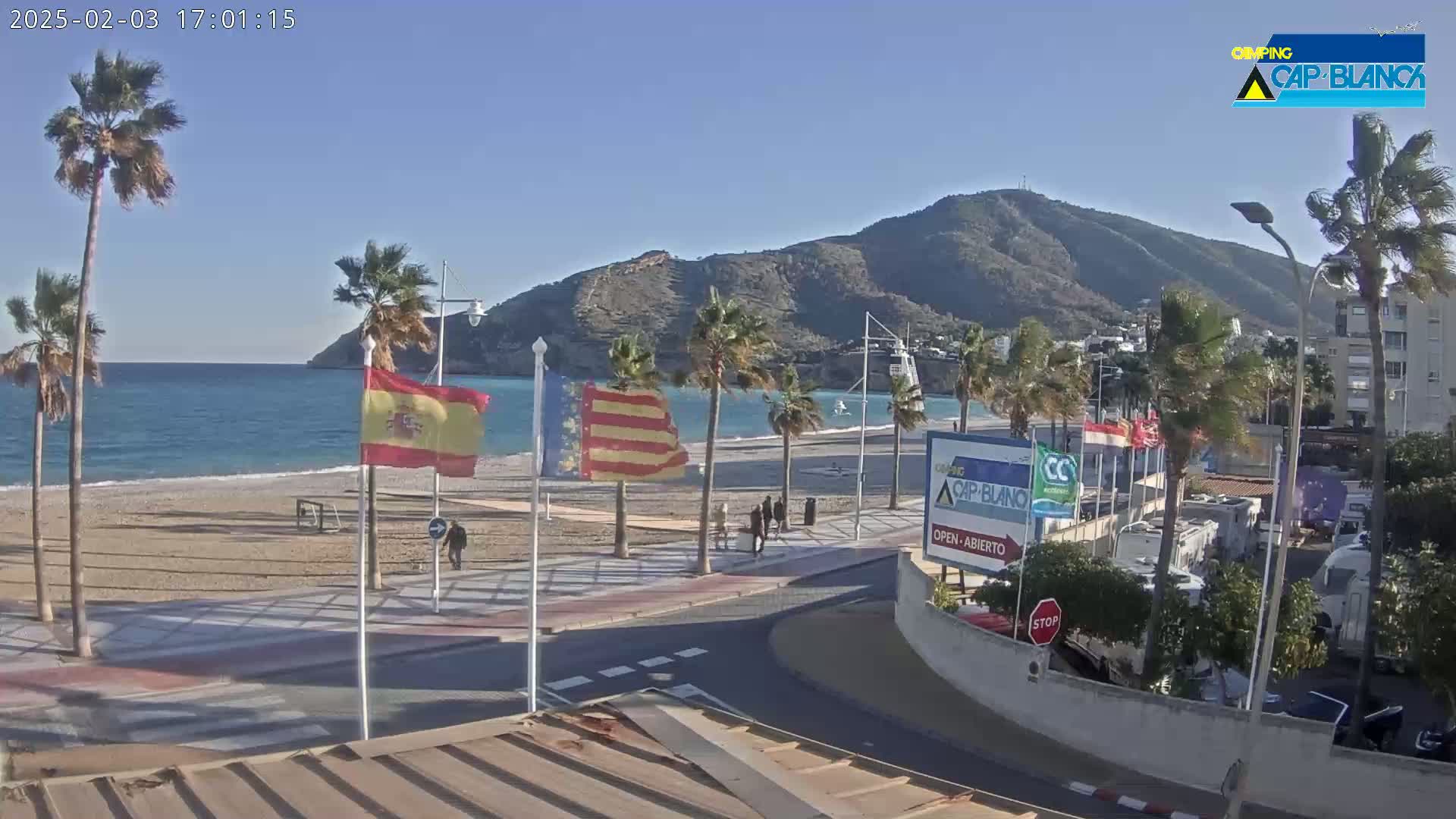 A sandy beach with palm trees and a calm ocean is situated next to a paved walkway and a parking lot with RVs under a clear, sunny sky and a mountainous backdrop.