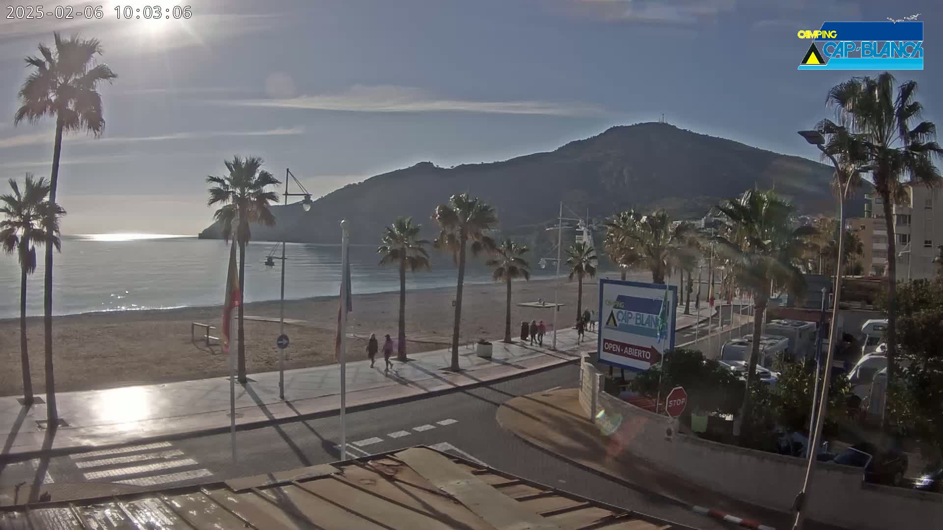 A sunny day reveals a beach scene with calm water, palm trees lining a promenade, and a mountain in the background.