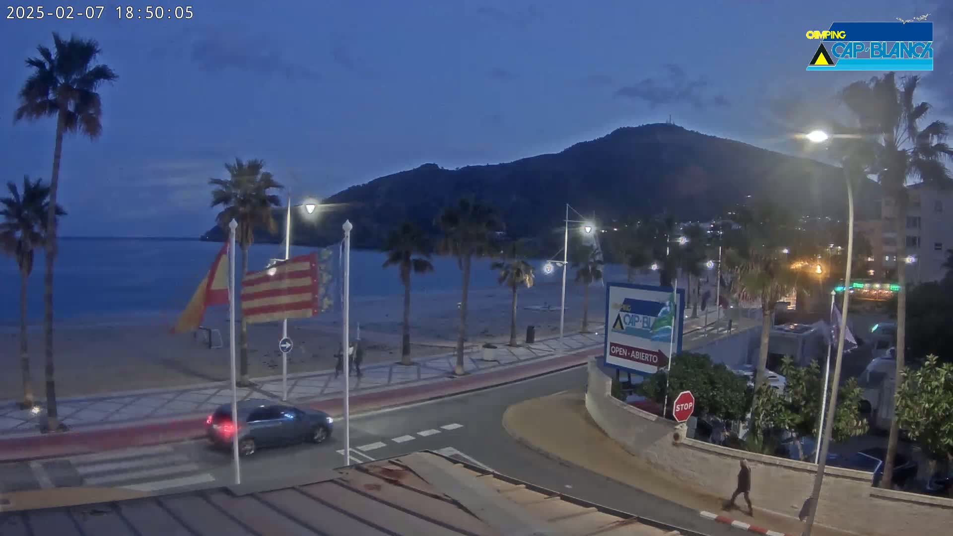 A twilight beach scene shows palm trees lining a paved walkway next to a sandy beach and calm ocean, with a hill in the background under a clear, dark blue sky.