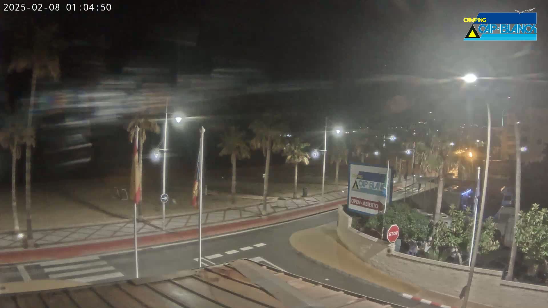A nighttime view of a beachfront road with palm trees, streetlights, and a sandy beach visible in the background under a partly cloudy sky.