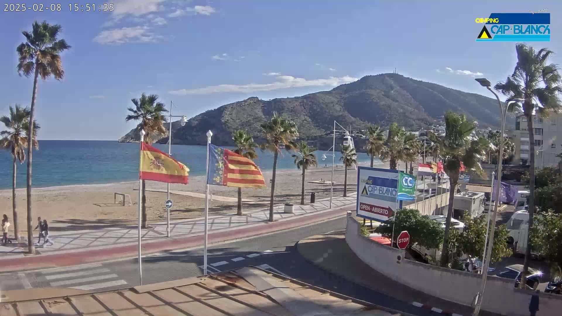A sunny beach scene shows a sandy beach, calm ocean, palm trees, and a mountain in the background, with a roadway and buildings visible in the foreground.