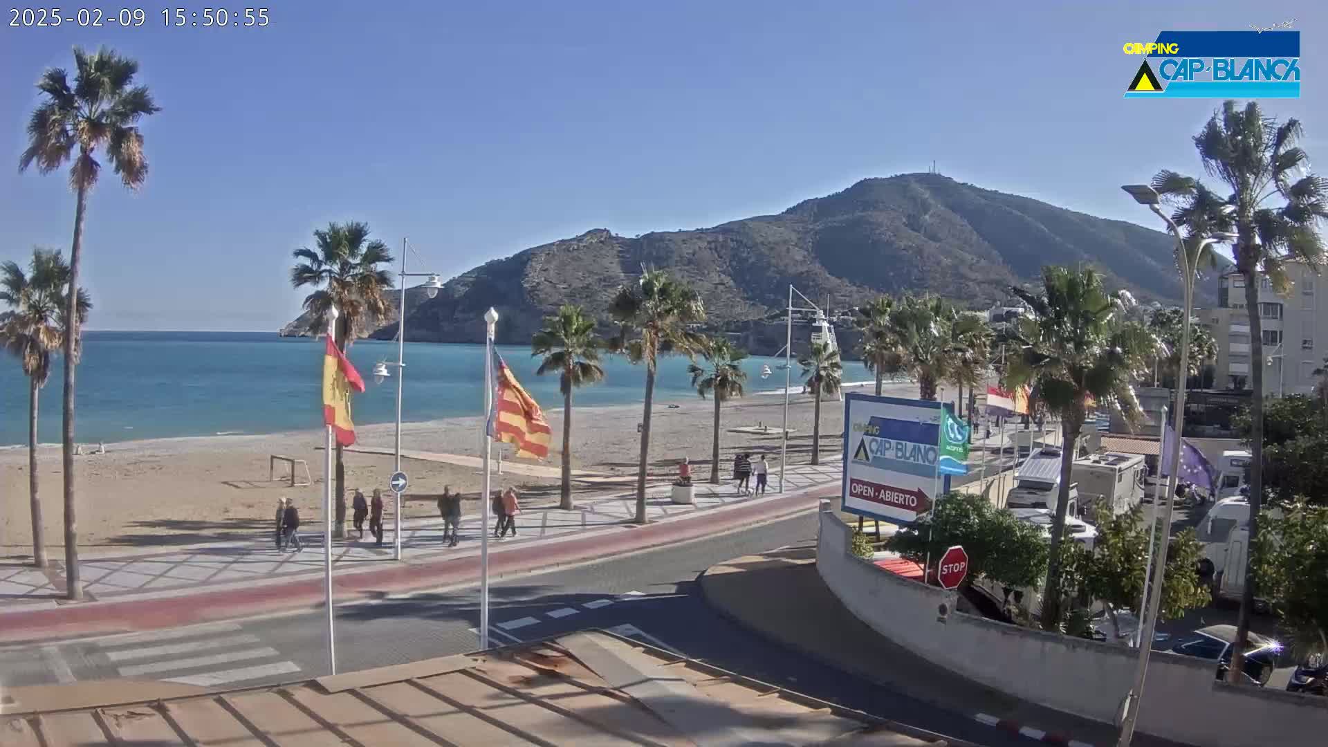 A sunny beach scene shows a sandy shoreline, calm blue water, palm trees lining a paved walkway, and a few people walking near a mountain backdrop.