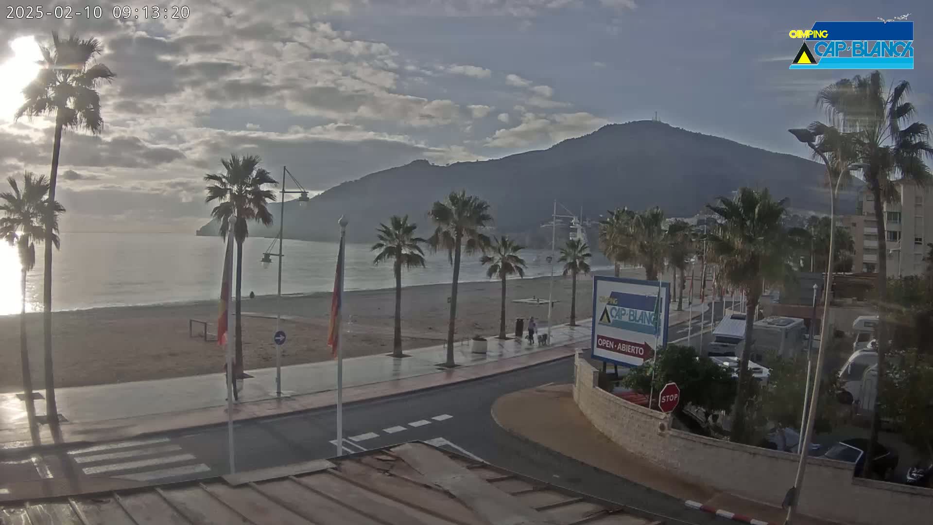 A mostly cloudy morning shows a sandy beach with palm trees lining a paved walkway next to a calm sea and a mountain in the background.