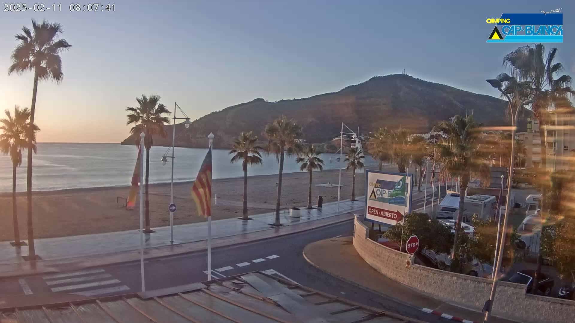 A sunrise view of a beach with palm trees lining a paved walkway next to a calm sea and a hill in the background.