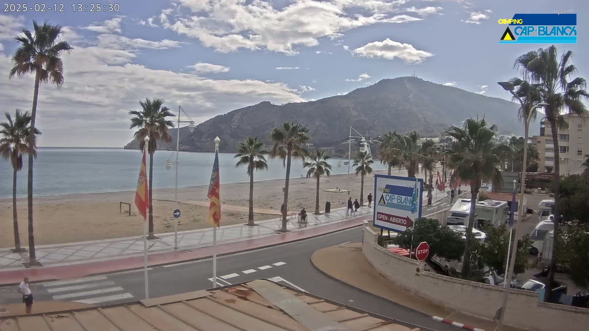 A partly cloudy day at a beachside promenade features palm trees, a sandy beach, calm water, a mountain in the background, and several people walking along the walkway.