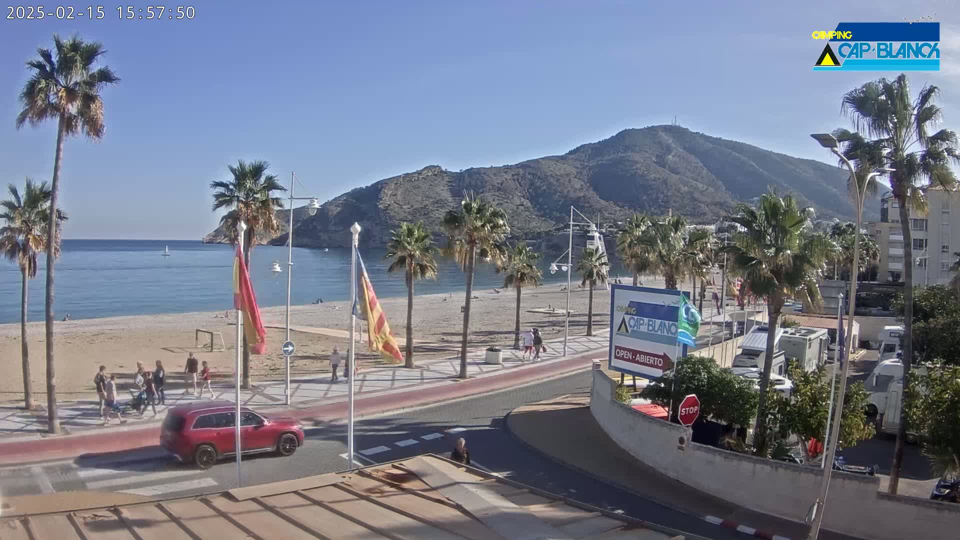 A sunny beach scene shows a sandy shore, calm blue water, palm trees lining a paved walkway, a red SUV, and a mountain in the background.