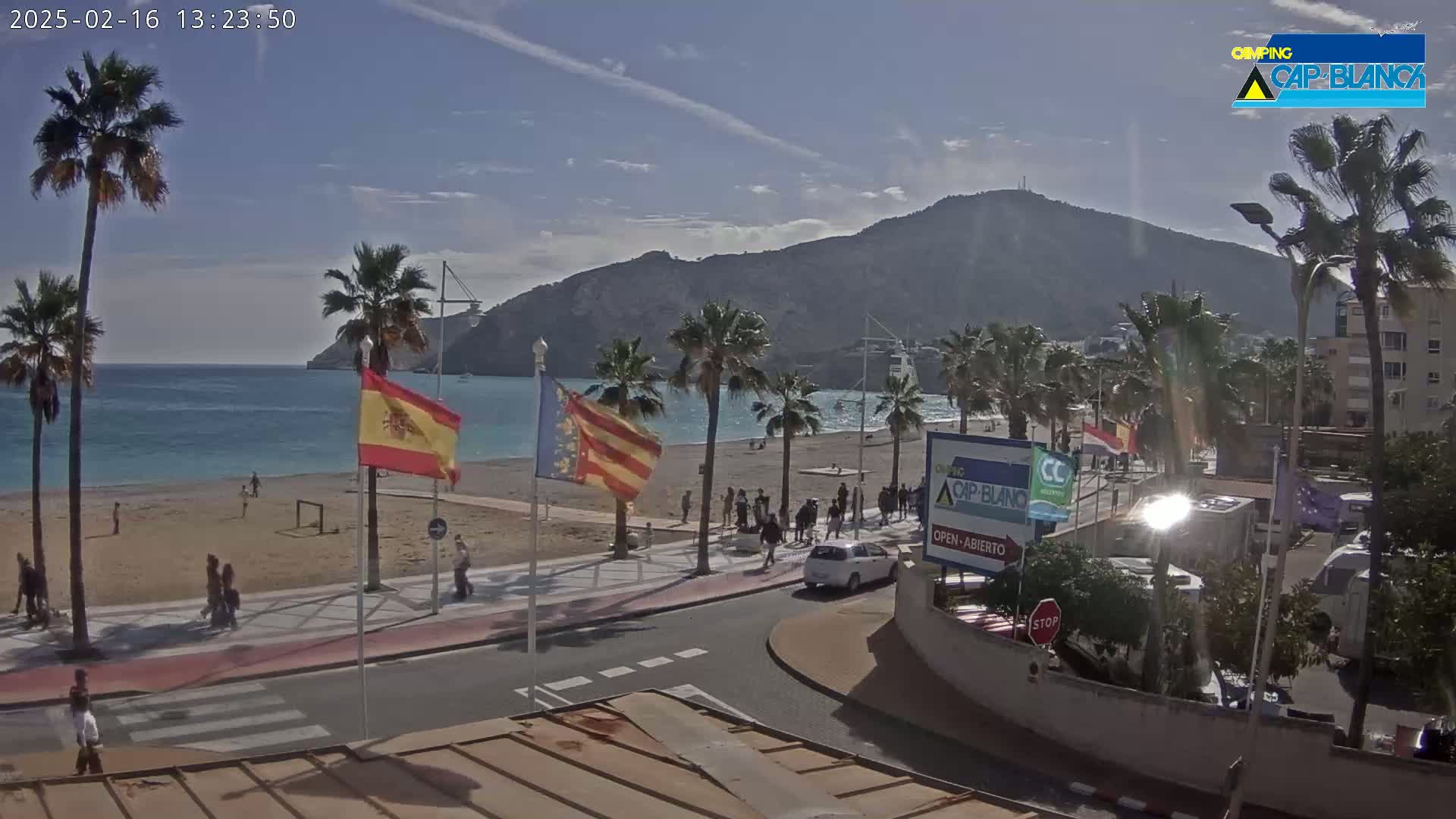 A mostly sunny beach scene shows a sandy beach with people walking along the shore and a boardwalk lined with palm trees, overlooking a calm ocean with a mountain in the background.