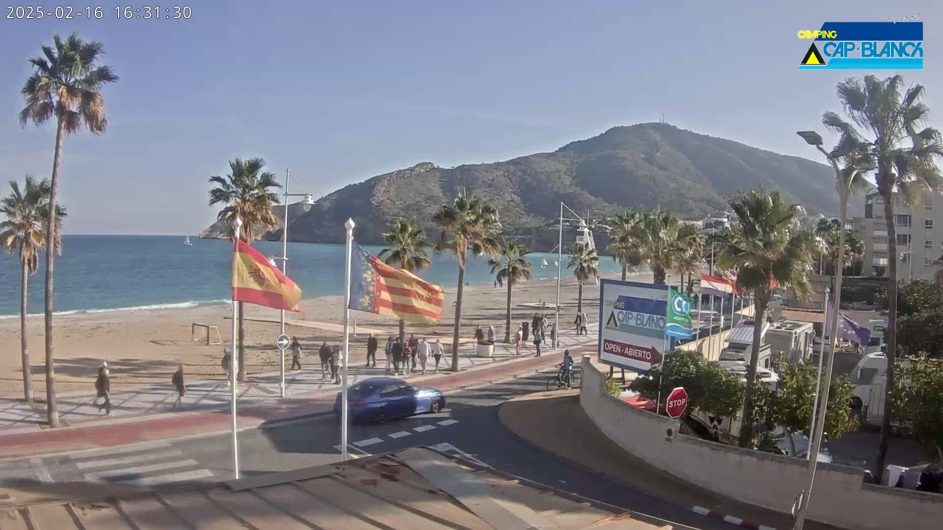 A sunny beach scene shows a sandy shore with palm trees, people walking along a promenade, a few cars parked, and mountains in the background.