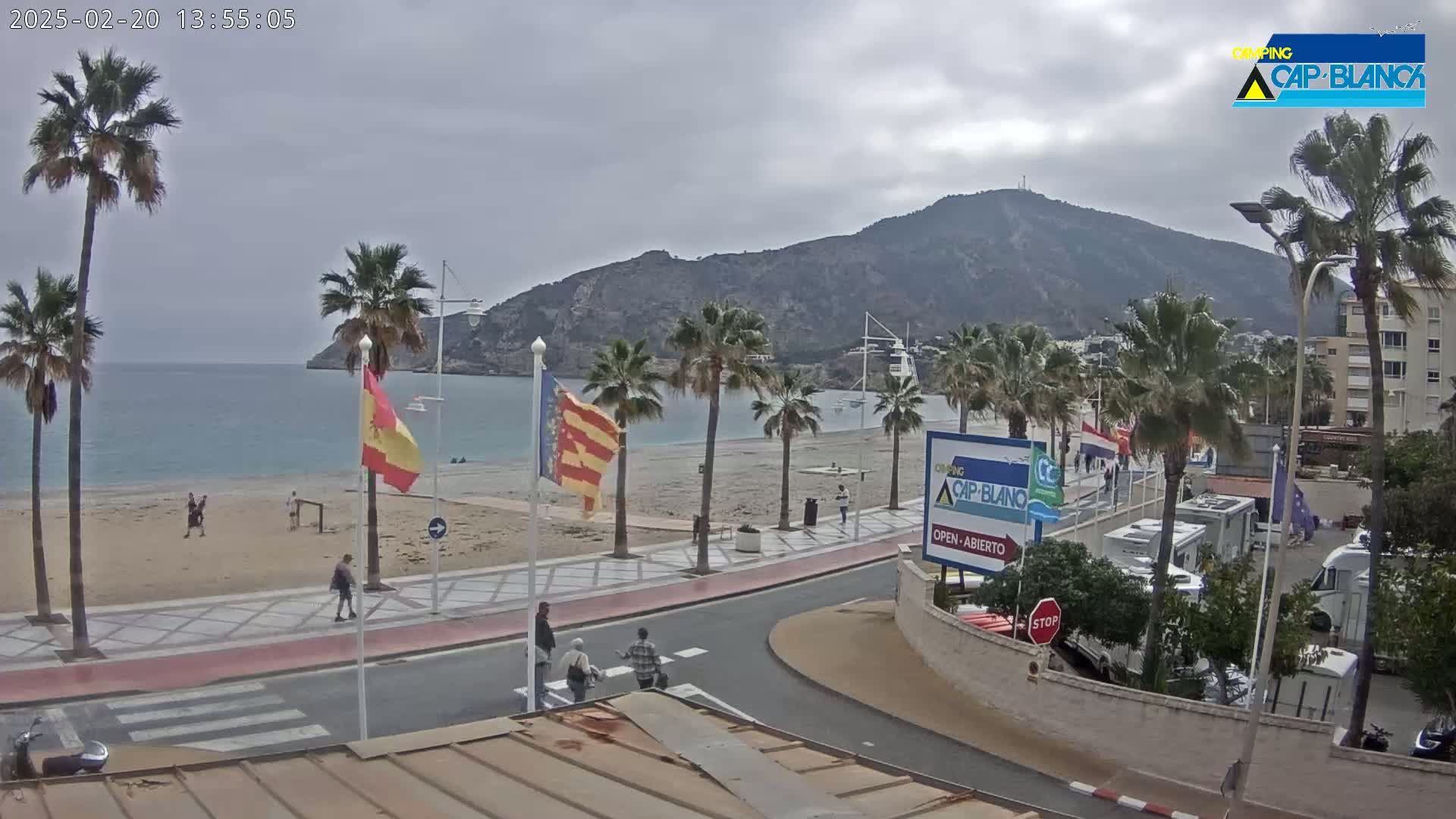 A sandy beach with palm trees lines a paved walkway next to a calm bay, under an overcast sky, with a mountain in the background.