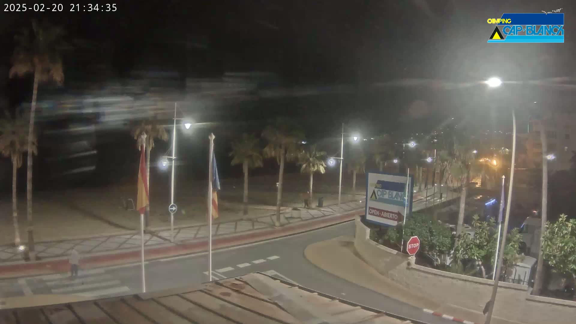 A nighttime view of a beachside promenade with palm trees, streetlights, and a few people walking under a slightly cloudy sky.