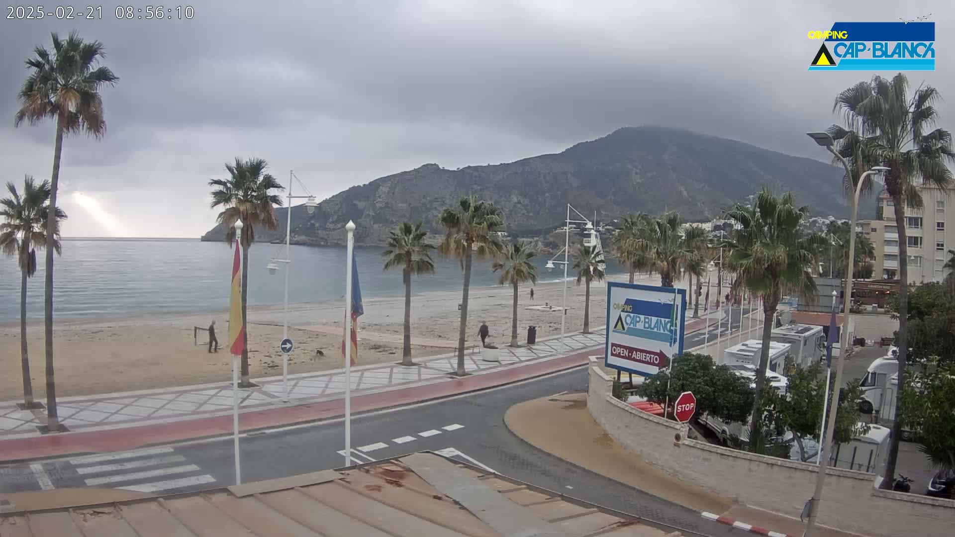 A mostly cloudy day shows a sandy beach with palm trees lining a paved walkway next to a calm ocean, with a mountain in the background and RVs parked nearby.