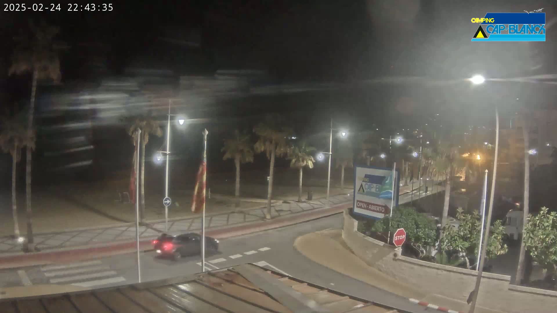 A nighttime street scene shows a car driving past palm trees lining a beachfront road under hazy conditions.