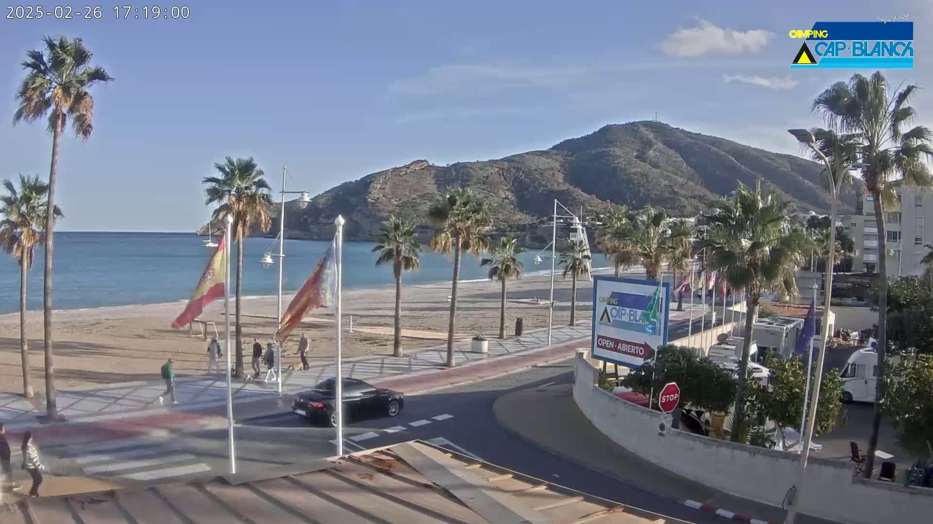 A sandy beach with palm trees and a mountain in the background on a sunny day, with a few people walking along the promenade and cars driving on the road.