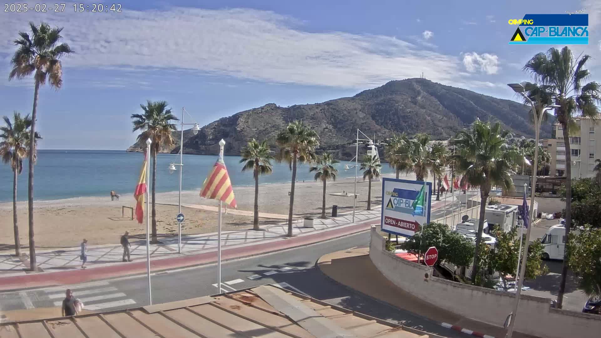 A sandy beach with palm trees lines a paved road leading to a seaside town nestled against a mountain, under a partly sunny sky.