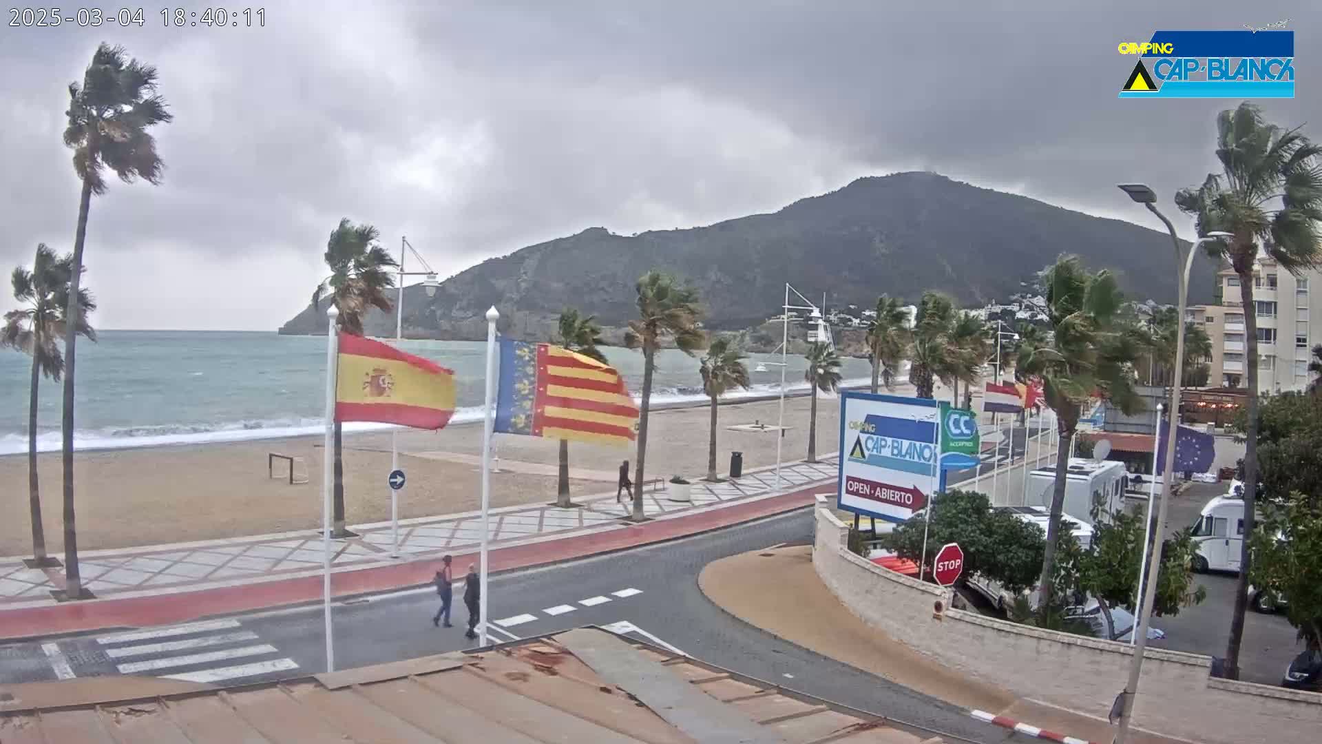 A windy, overcast day at a beachside promenade shows palm trees bending in the wind, a choppy sea, and a few people walking along the path near parked RVs.