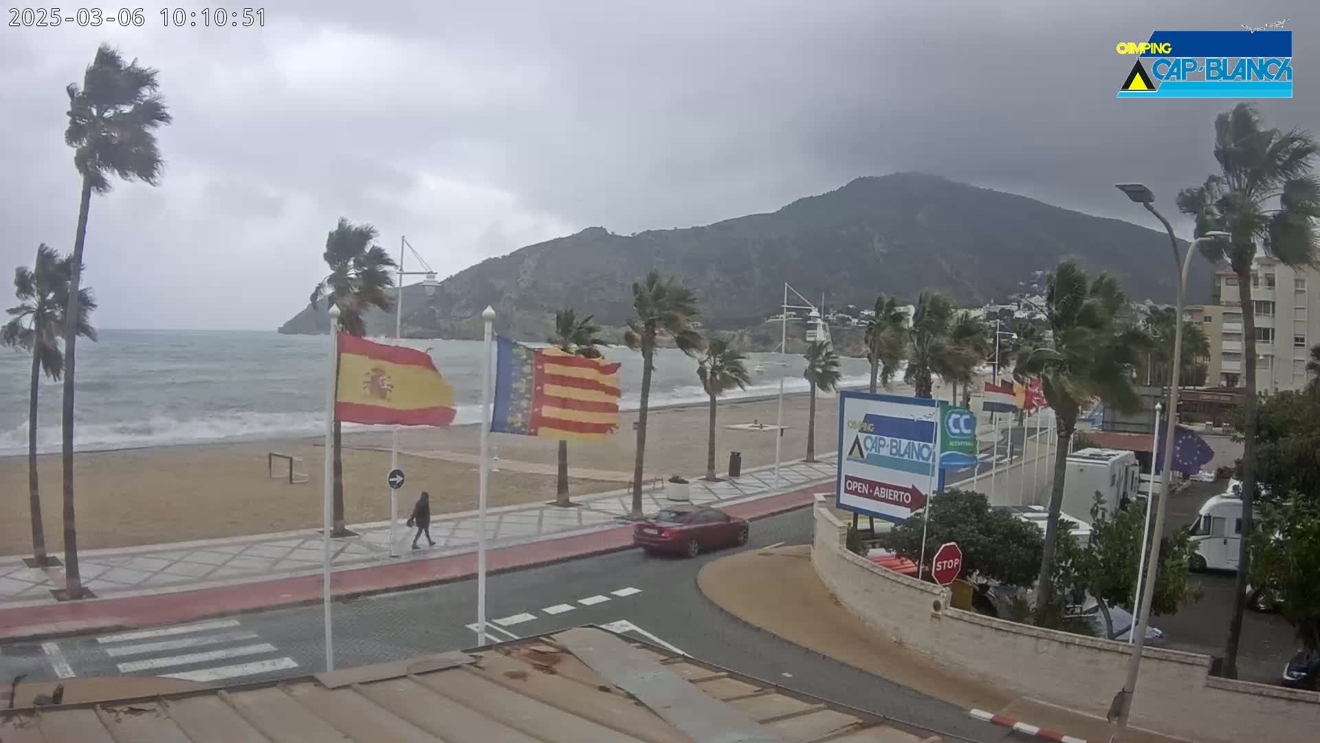 A windy, overcast day at a beachside promenade shows palm trees bending in the wind, choppy waves, and a few people walking near parked cars and RVs.