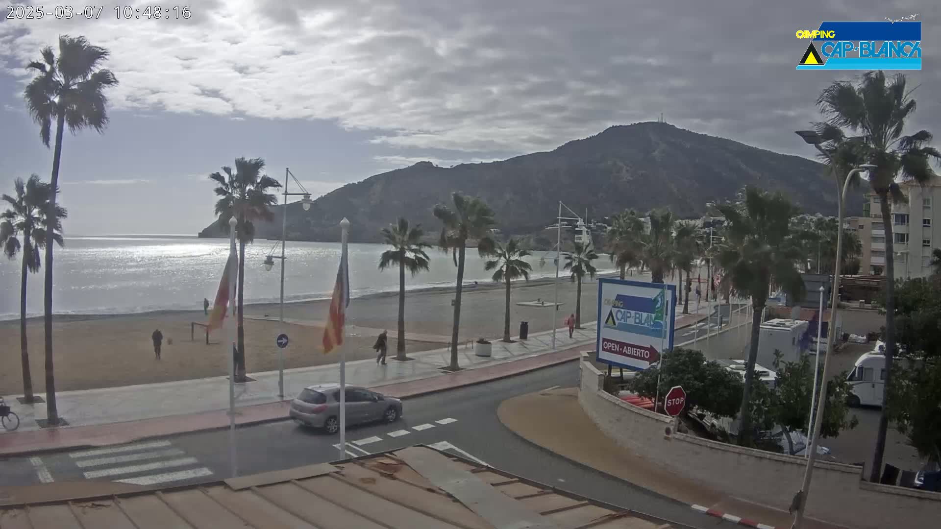 A partly cloudy day shows a beach with palm trees lining a paved walkway next to a calm sea and a mountain in the background.