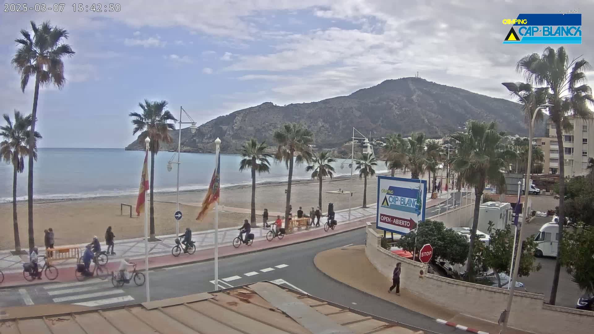 A mostly sunny day shows a sandy beach with palm trees lining a paved walkway next to calm blue ocean water, a mountain in the background, and several people riding bicycles and walking.