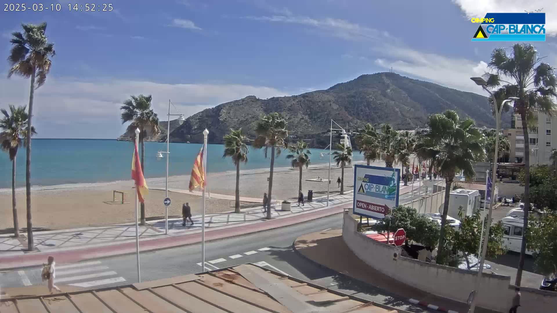A sandy beach with palm trees and a calm, turquoise sea stretches along a paved walkway and road beneath a mostly sunny sky, with a mountain in the background.