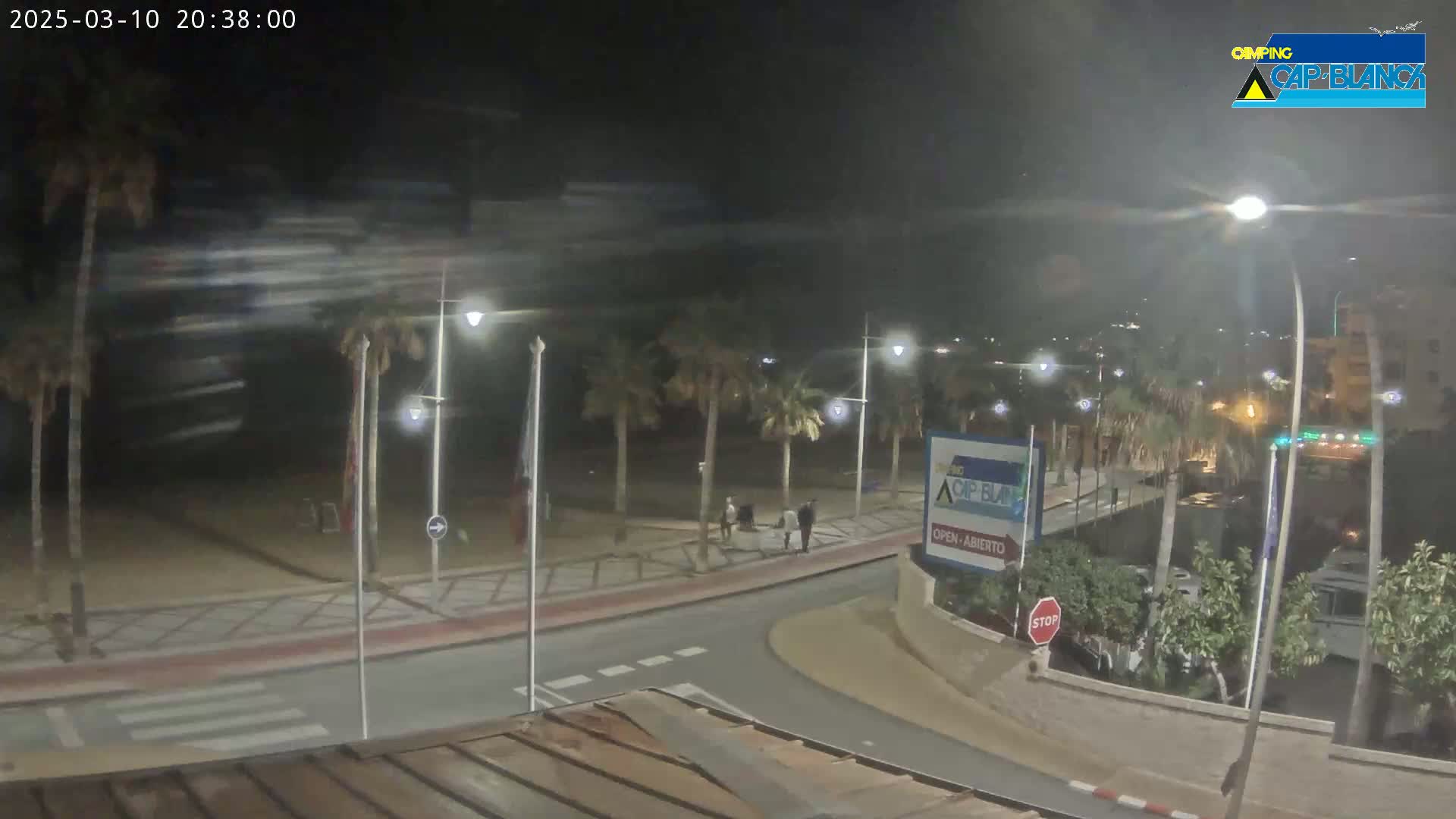 A nighttime view of a palm tree-lined street and beach, with a few people walking along the promenade under somewhat cloudy skies.