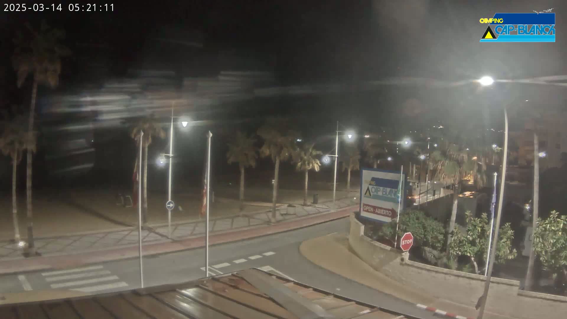 A nighttime view of a palm tree-lined road next to a beach, under cloudy skies.