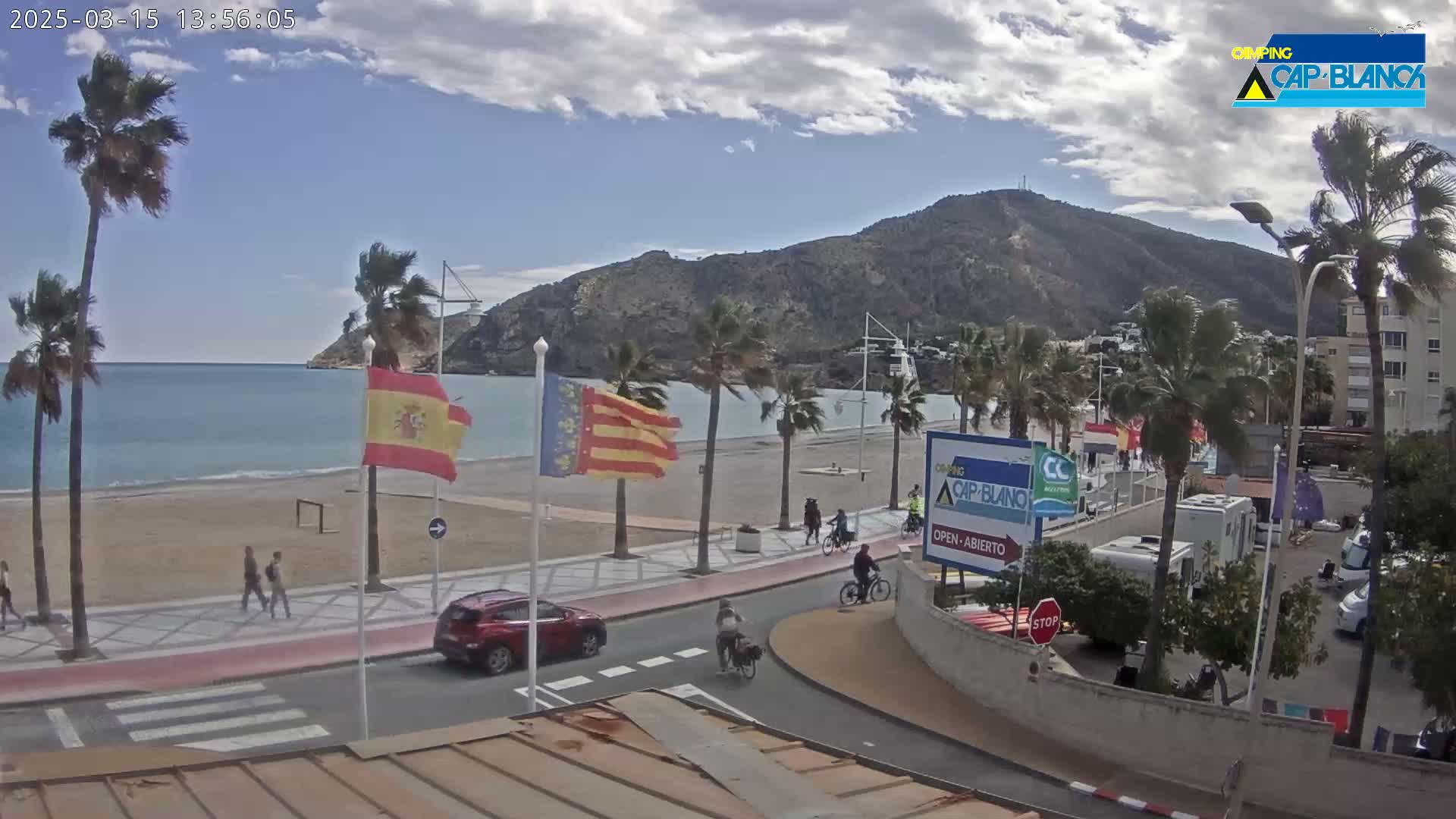 A partly cloudy day shows a beach with palm trees, a road with people and vehicles, and a mountain in the background.