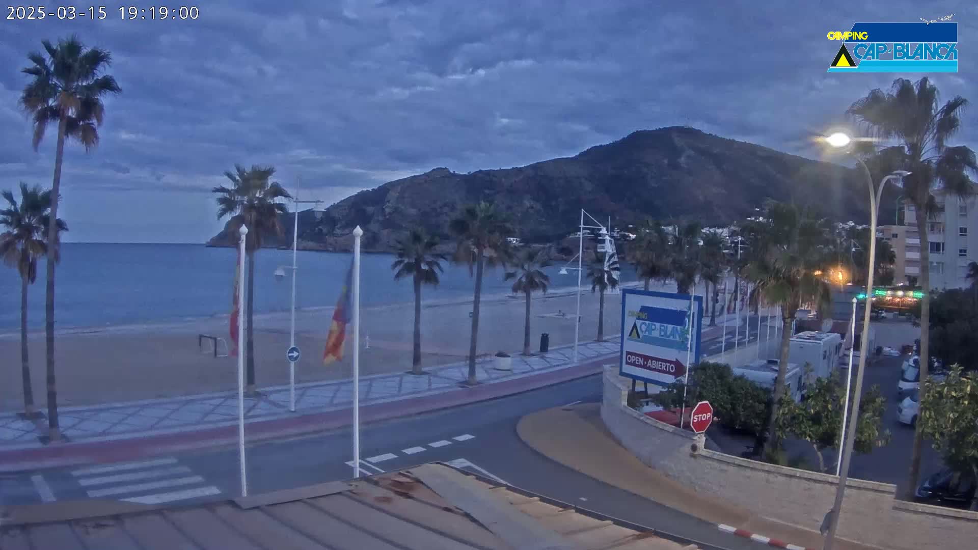 A sandy beach with palm trees and a calm sea is seen under a cloudy twilight sky, with a hill in the background and a road leading to buildings and parked vehicles.